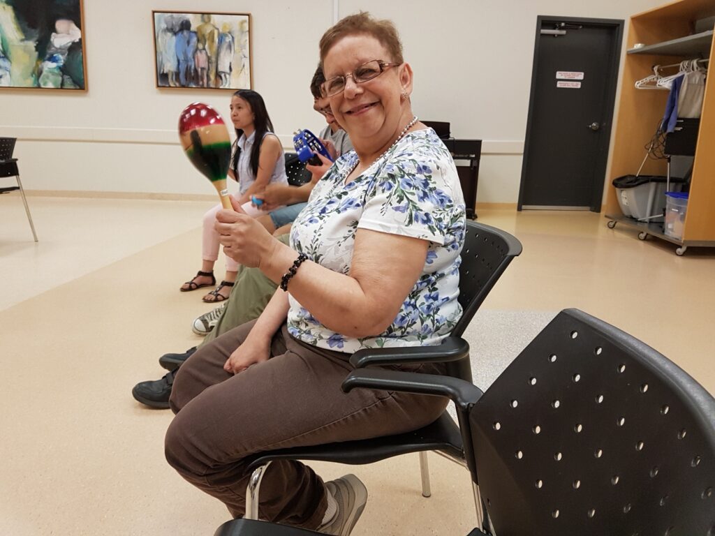 A smiling person in a blue patterned floral shirt shaking a wooden maraca while sitting in a group circle