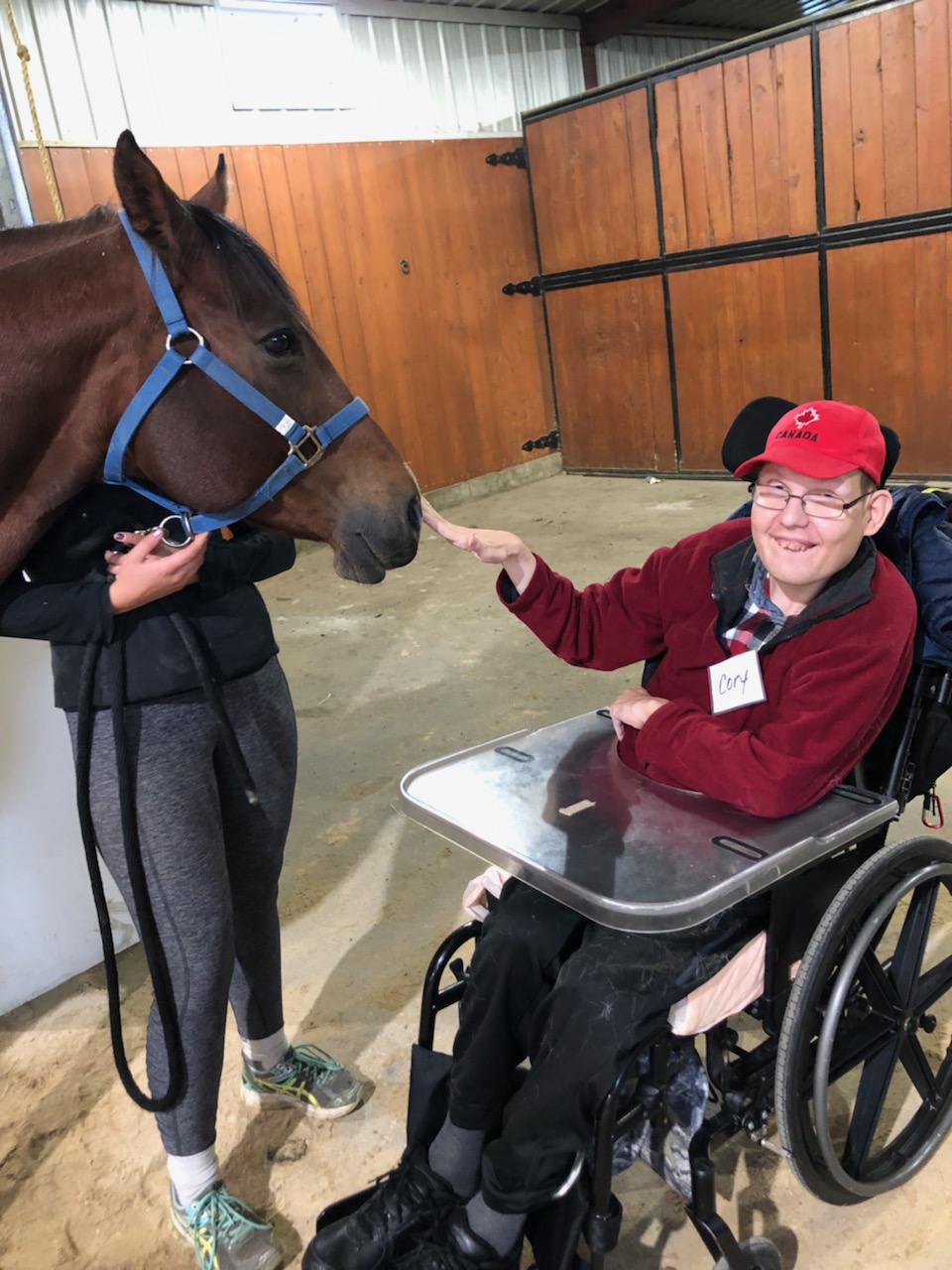 A happy resident in a wheelchair with a name tag that reads Cory. Cory is inside of a barn and petting a horse on the nose.