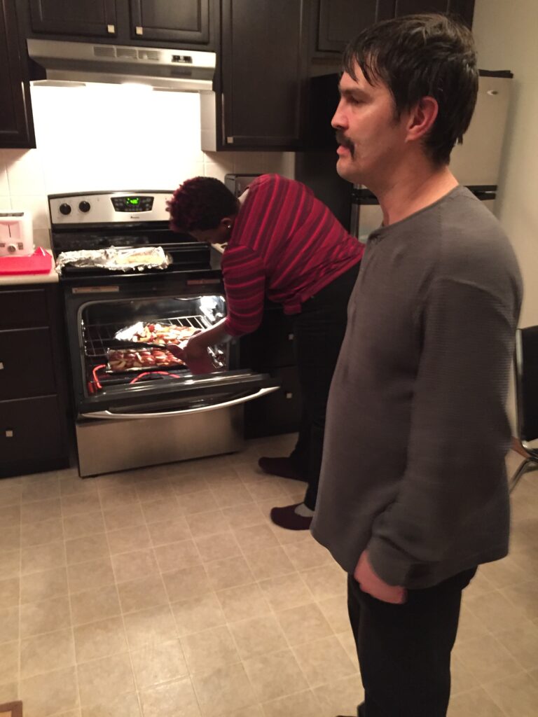 A person standing in the middle of the kitchen while another person puts a tray of vegetables into the oven