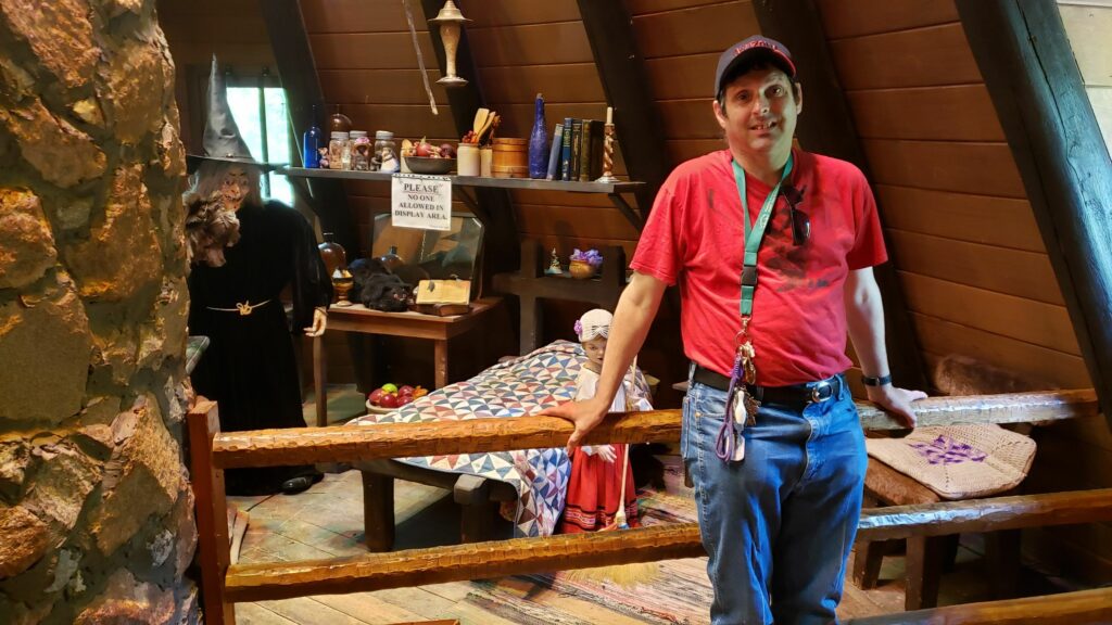 A resident in a black baseball cap and a bright red t-shirt leaning against a fence inside of the Witch's Hut in Kildonan Park